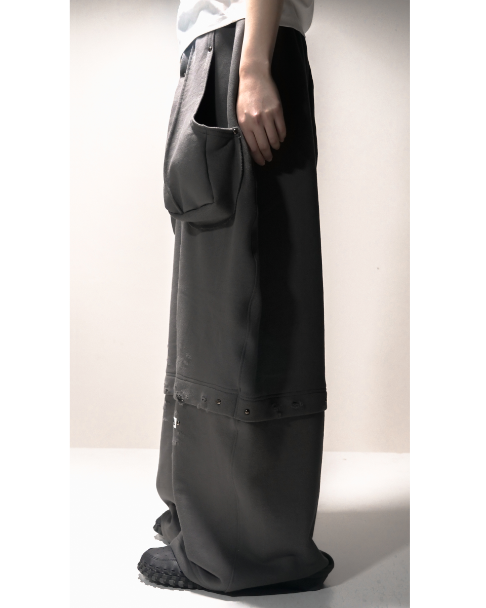 모델이 진회색 와이드핏 스웨트 팬츠를 입은 다른 측면 모습. 옆 포켓이 크게 부착되어 있고, 무릎 아래 부분에는 해진 원단과 스터드 장식이 반복되어 있다.