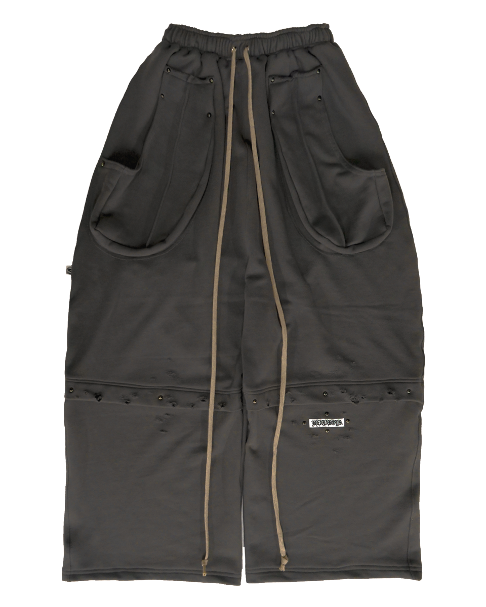 진회색 와이드핏 스웨트 팬츠 정면. 허리 밴딩에 긴 베이지색 끈이 두 줄 늘어져 있고, 양옆에는 큰 입체 포켓이 달려 있다. 무릎 아래 부분에는 찢김과 구멍 디테일이 있으며, 검은색 스터드 장식이 줄지어 있고, 오른쪽 다리 아래에 흰색 라벨이 부착되어 있다.