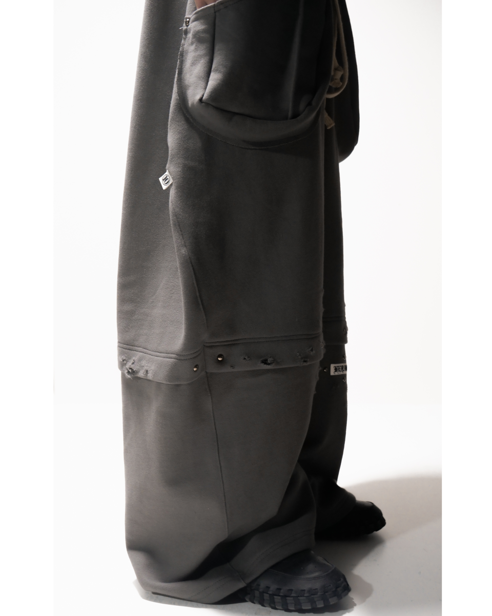 모델이 진회색 와이드핏 스웨트 팬츠를 입은 측면 모습. 바지 옆면에 큰 포켓이 튀어나와 있으며, 무릎 아래 부분에는 찢김 디테일과 스터드 장식이 한 줄로 이어져 있다. 전체적으로 바지통이 넓고 길다.”