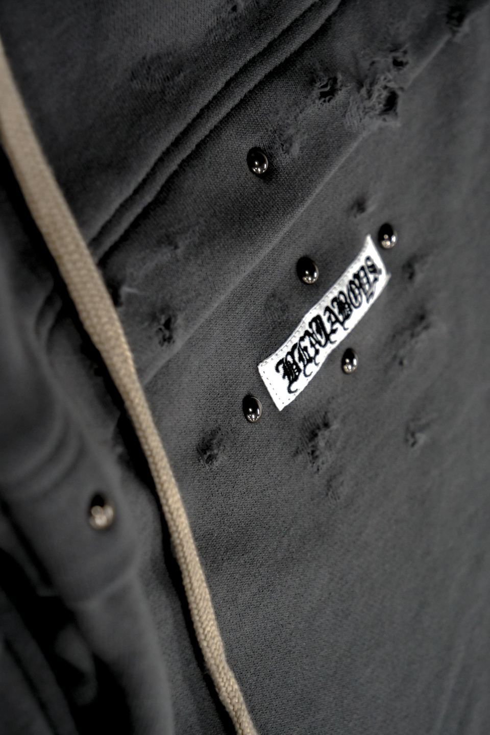 진회색 와이드핏 스웨트 팬츠 확대 사진. 원단에는 해진 디테일과 검은색 스터드 장식이 보이고, 흰색 라벨에는 ‘BENJABOYS’ 로고가 새겨져 있다. 옆에는 베이지색 끈이 함께 보인다.